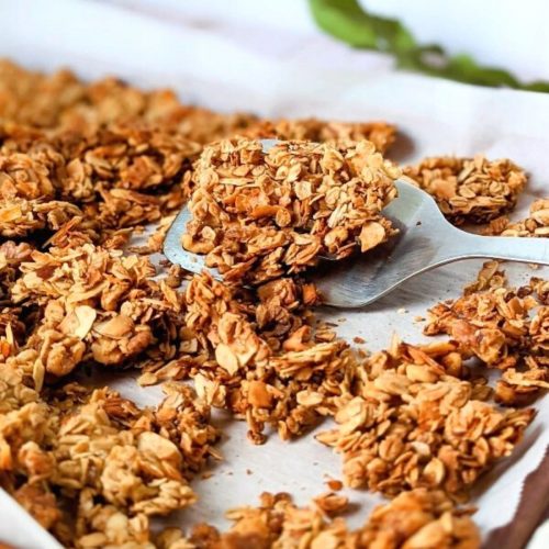 unsalted granola in a pan with oats maple syrup coconut walnuts almonds vanilla being scooped by a spatula on a sheetpan.
