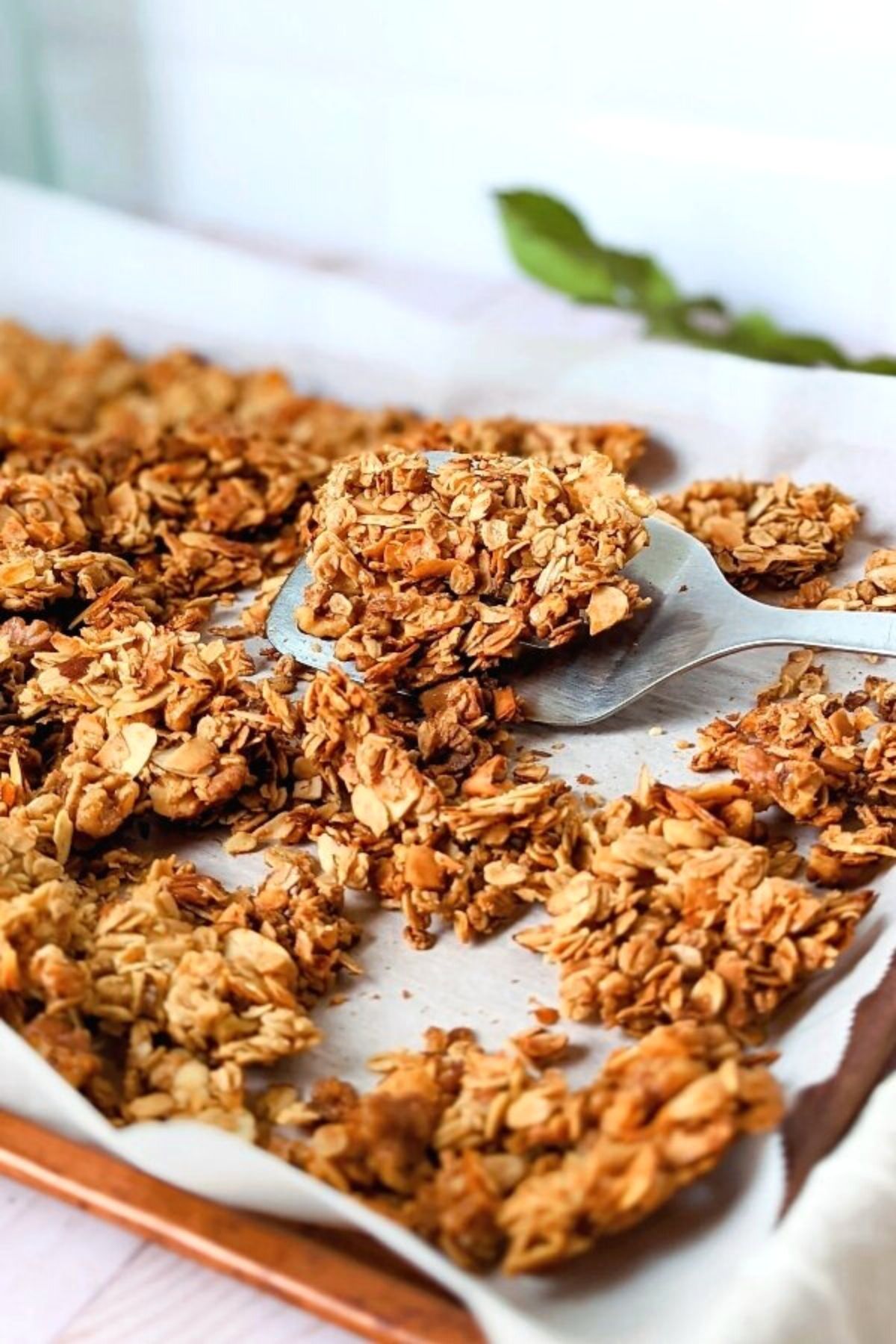 unsalted granola in a pan with oats maple syrup coconut walnuts almonds vanilla being scooped by a spatula on a sheetpan.
