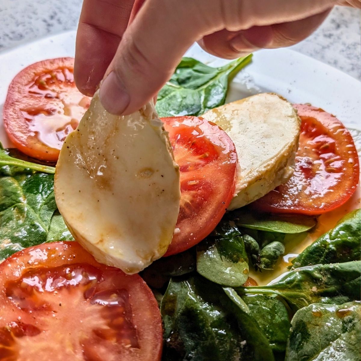 a hand placing a slice of mozzarella cheese on a plate with basil and tomatoes.