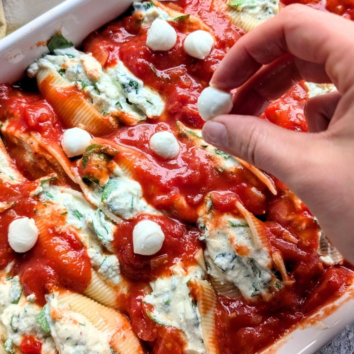 a hand sprinkling fresh mozzerella pearls on a baking dish of stuffed shells with low sodium tomato sauce