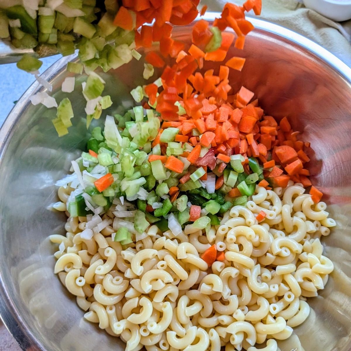 carrots, celery, onions, and bell peppers being added to cooked macaroni for salad.