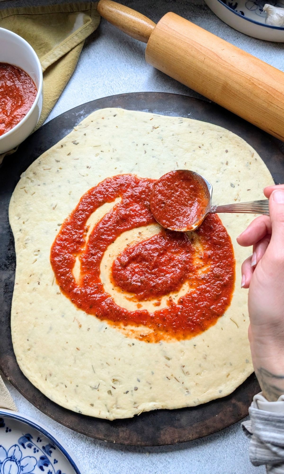 a hand spooning low salt pizza sauce on homemade low sodium crust with a rolling pin in the background