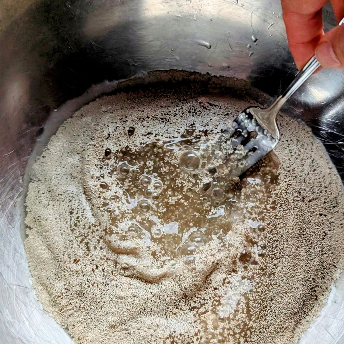 a bowl of warm water, yeast, and sugar being mixed with a fork to make pizza dough